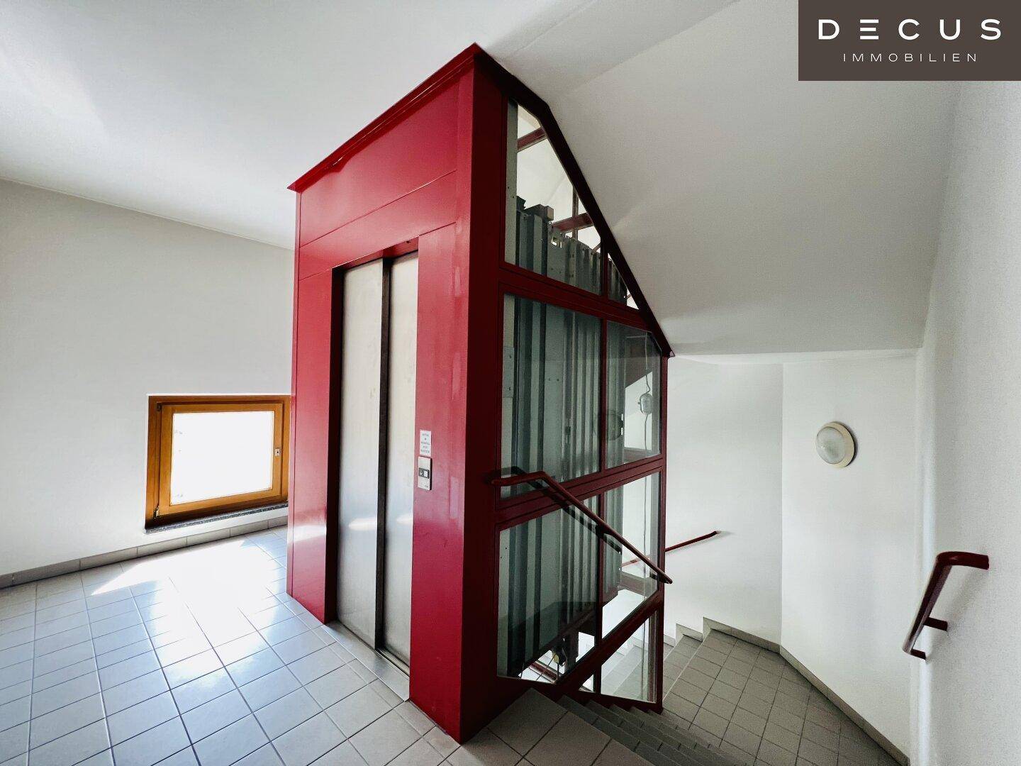Treppenhaus mit einem modernen roten Aufzug und Fenstern, die für Helligkeit sorgen.