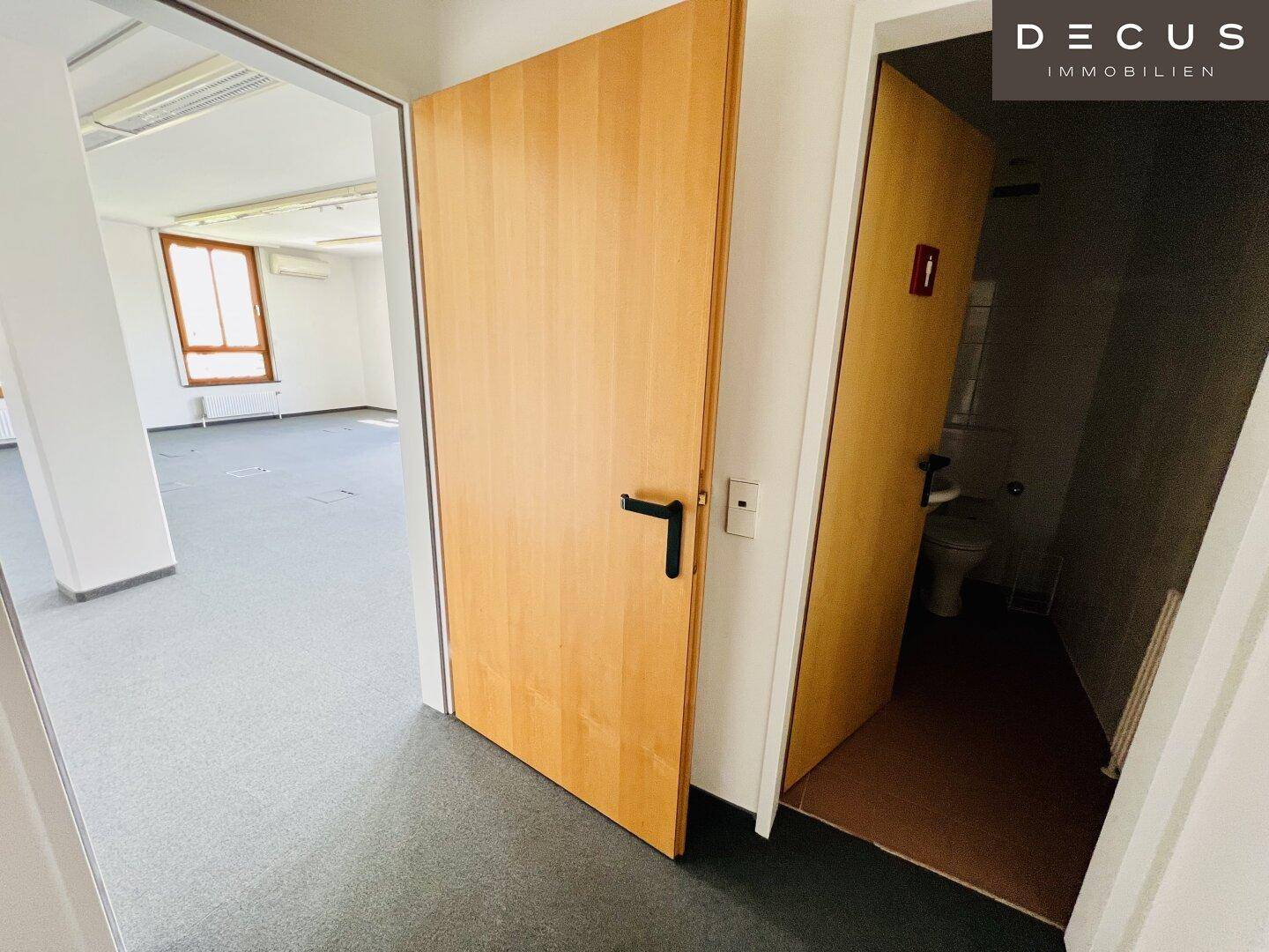 Blick vom Flur in eine Toilettenkabine und einen leeren Büroraum mit dunklem Teppichboden.