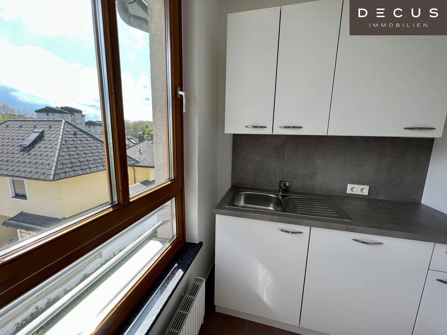Kleine Küchenzeile mit weißen Schränken, Spülbecken und einem Fenster mit Blick nach draußen.