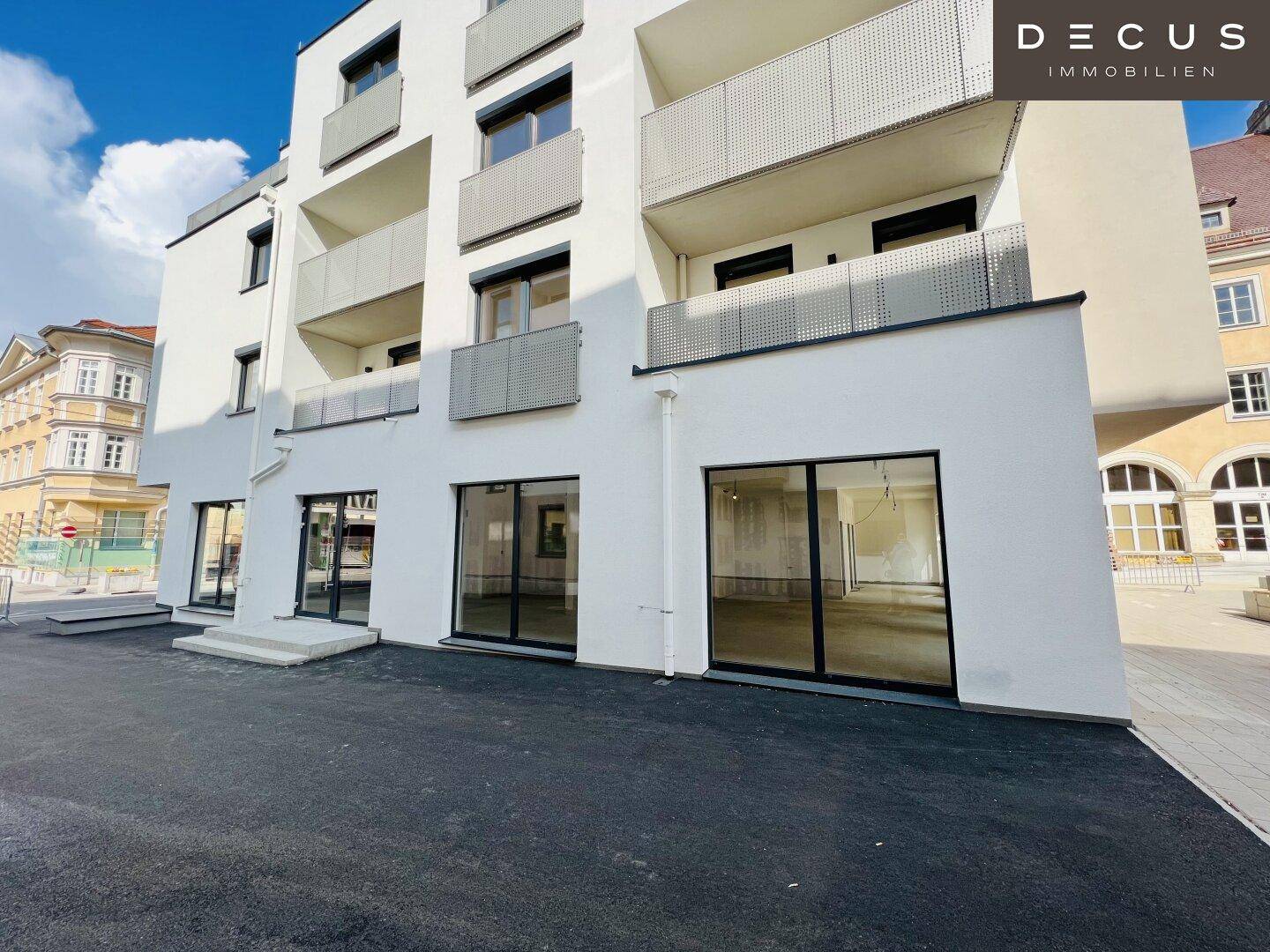 Moderne weiße Gebäudefassade mit Balkonen und großen Schaufenstern im Erdgeschoss, an einer Straße gelegen.