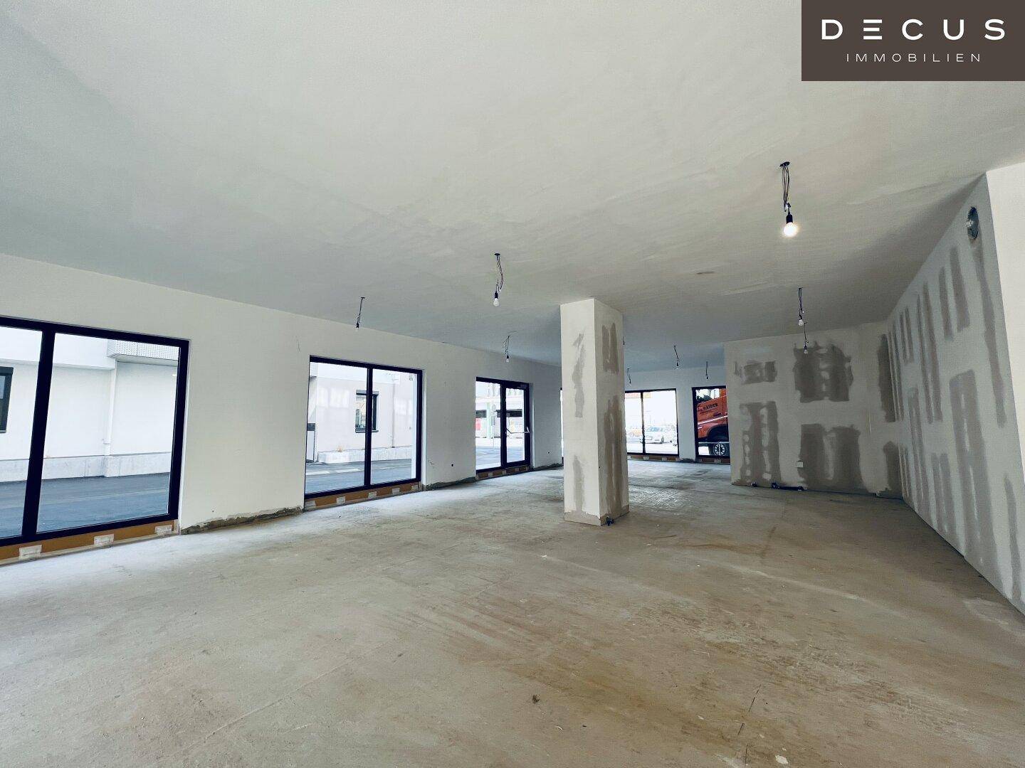 Geräumiger Gewerberaum im Rohbau mit Betonboden und vielen Fenstern, bereit für den Innenausbau.
