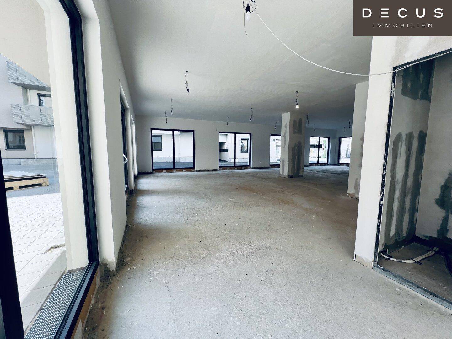 Heller Gewerberaum im Rohbau mit Betonboden und großen Fensterfronten, die den Raum belichten.
