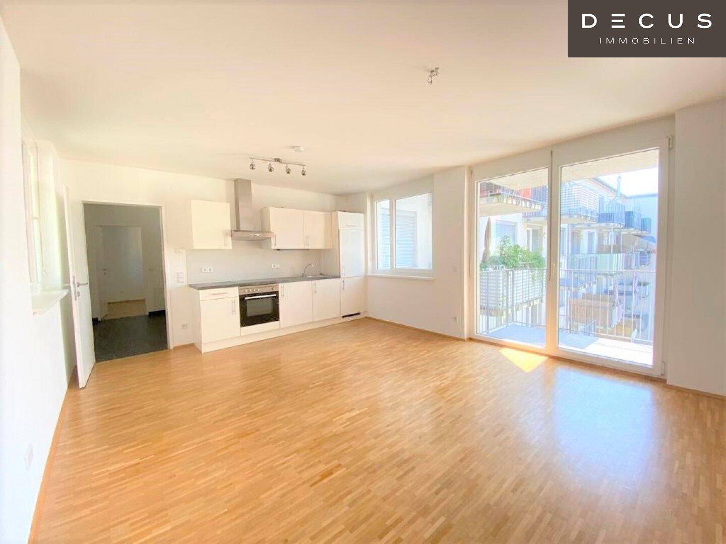 Heller offener Wohnbereich mit integrierter Küche, Parkettboden und Zugang zum Balkon.