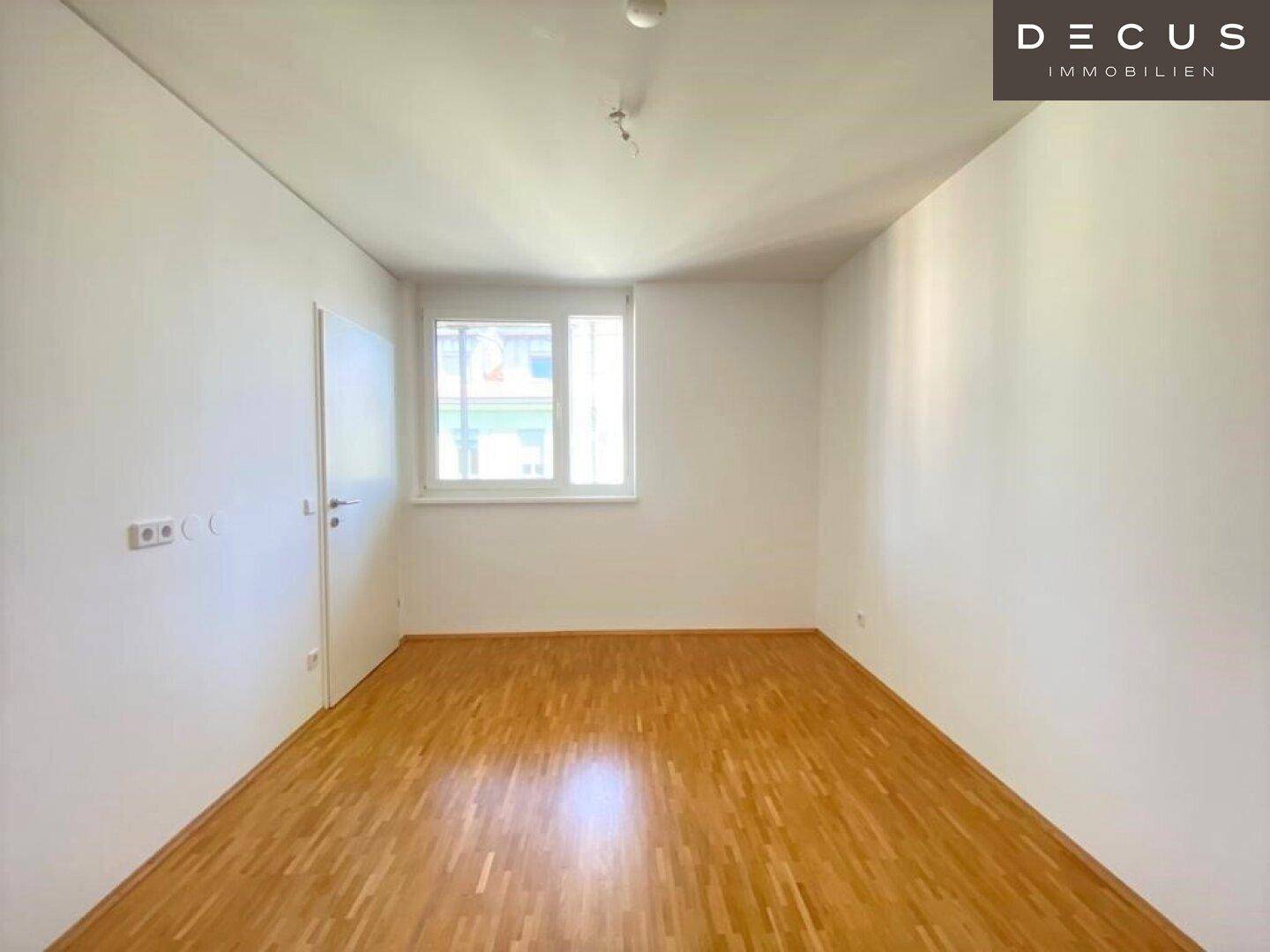 Leeres Schlafzimmer mit Parkettboden und einem Fenster, das viel Tageslicht hereinlässt.