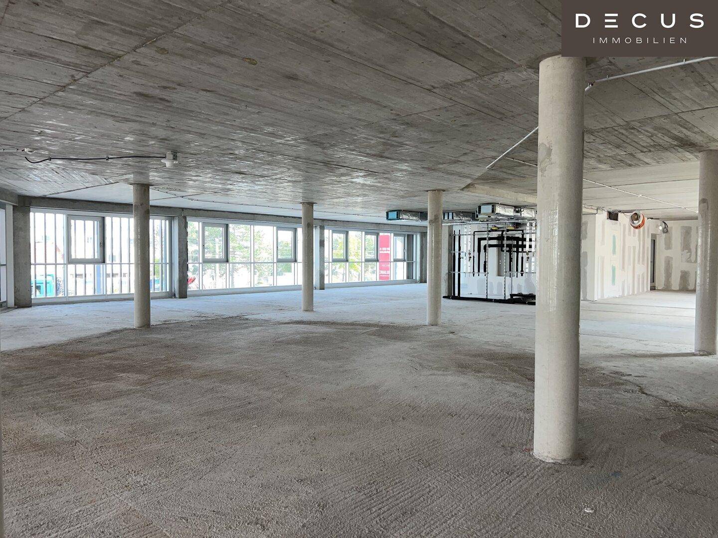 Weitläufiger, unmöblierter Bürobereich mit Betonsäulen, großen Fenstern und sichtbaren Installationen an der Decke.