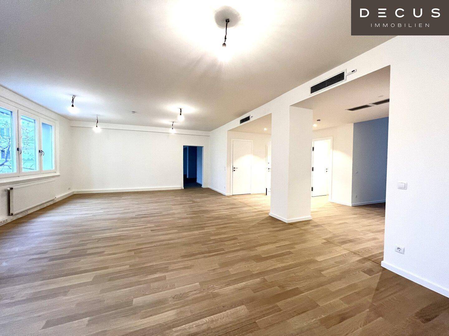 Geräumiger Wohnbereich mit Parkettboden und großen Fenstern, die viel Tageslicht hereinlassen.