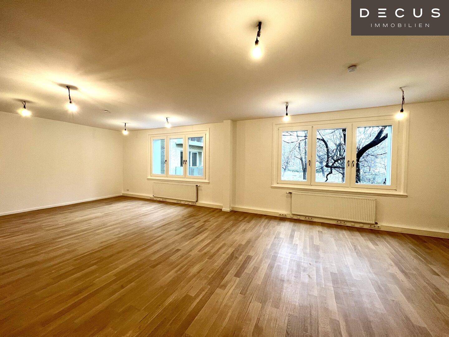 Heller Raum mit Holzboden und zwei Fenstern, die einen Blick auf Bäume nach draußen ermöglichen.