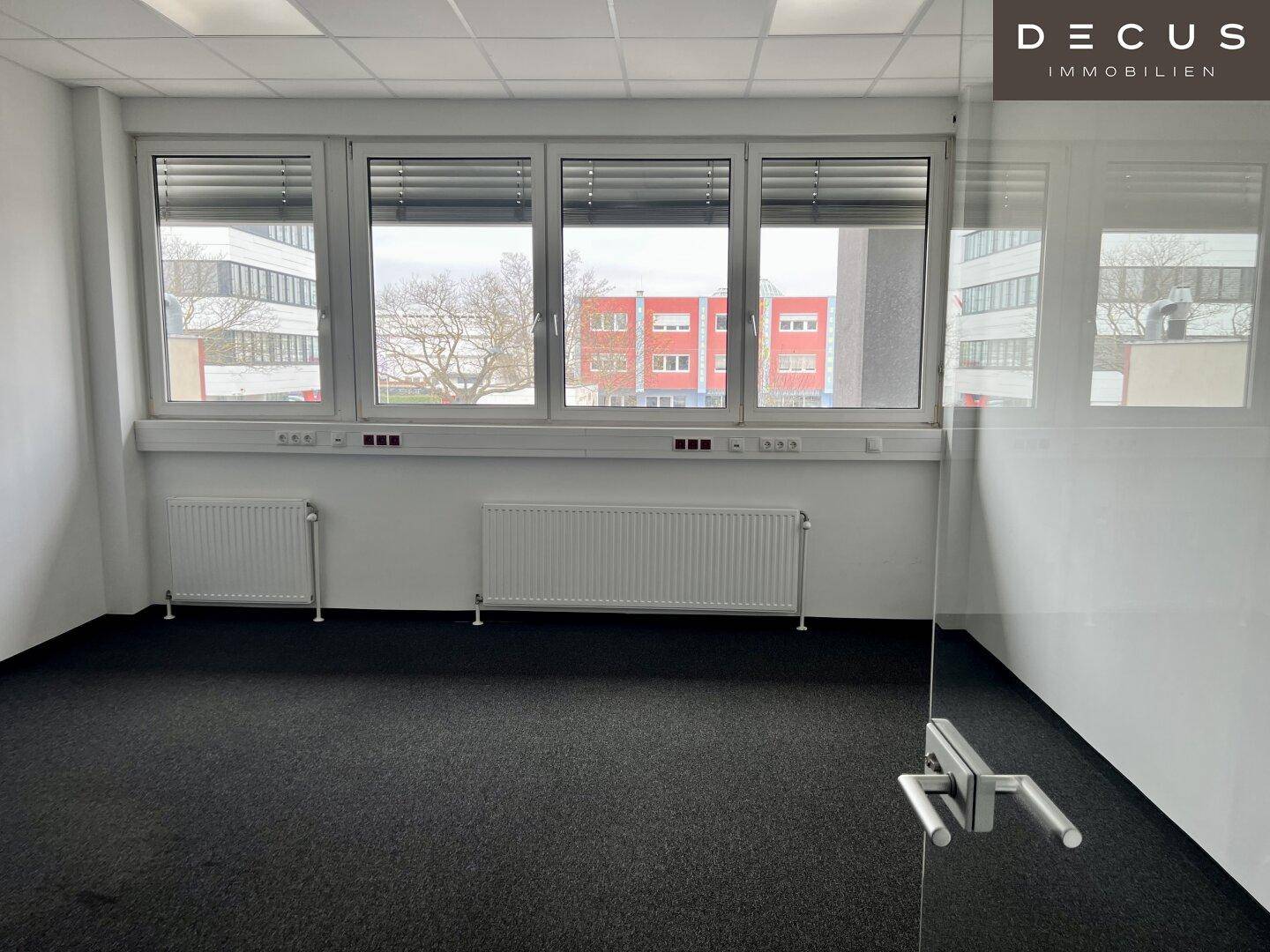 Geräumiges Büro mit Teppichboden, Heizkörpern unter den Fenstern und Blick auf umliegende Gebäude.