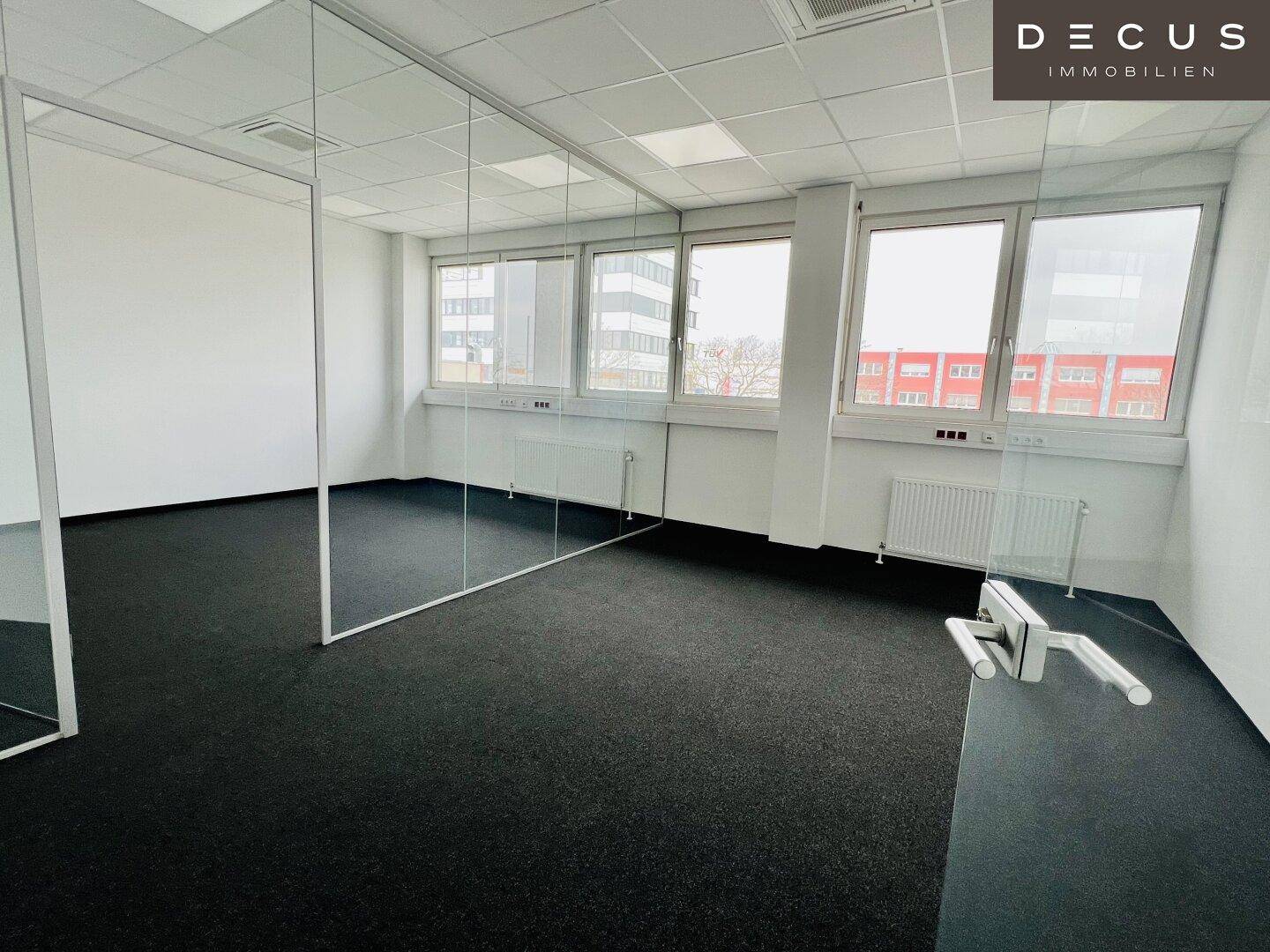 Helles Büro mit Teppichboden, großen Fenstern und modernen Glasabtrennungen, die den Raum flexibel gestalten.