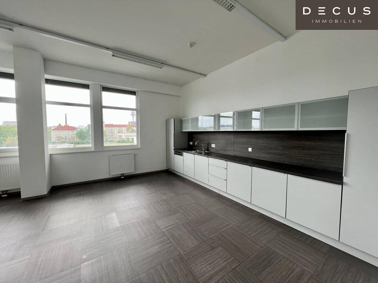 Moderne Teeküche in einem Bürogebäude mit weißen Schränken, dunkler Arbeitsplatte und großen Fenstern.