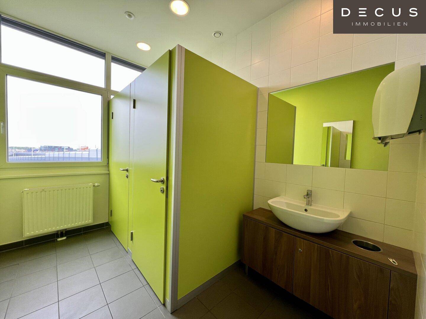 Helle Toilettenanlage mit grünen Kabinentüren, Waschbecken und Fenster für Tageslicht.