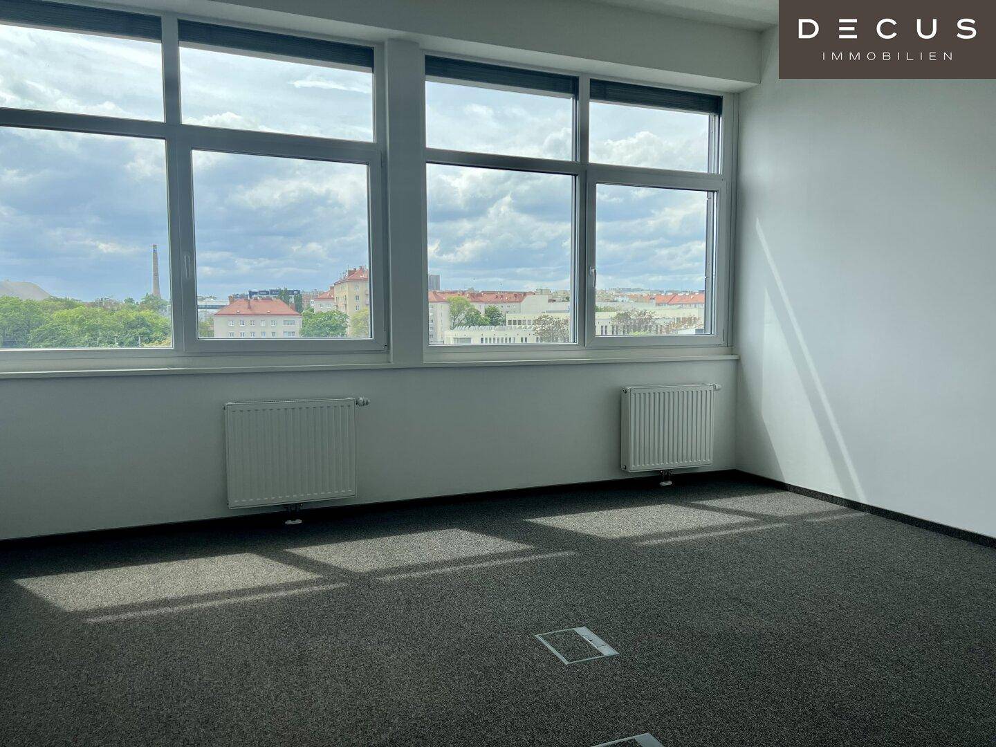 Heller Büroraum mit Teppichboden, großen Fenstern und Heizkörpern unter den Fenstern.