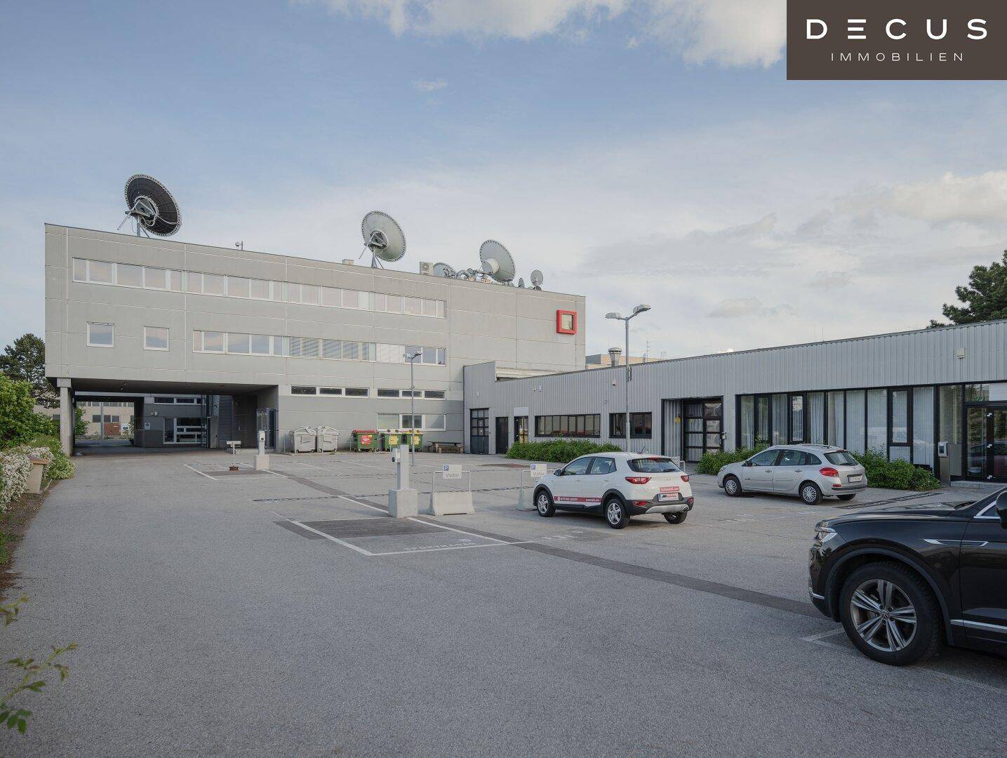 Gewerbegebäude mit großem Parkplatz und Satellitenschüsseln auf dem Dach unter bewölktem Himmel.