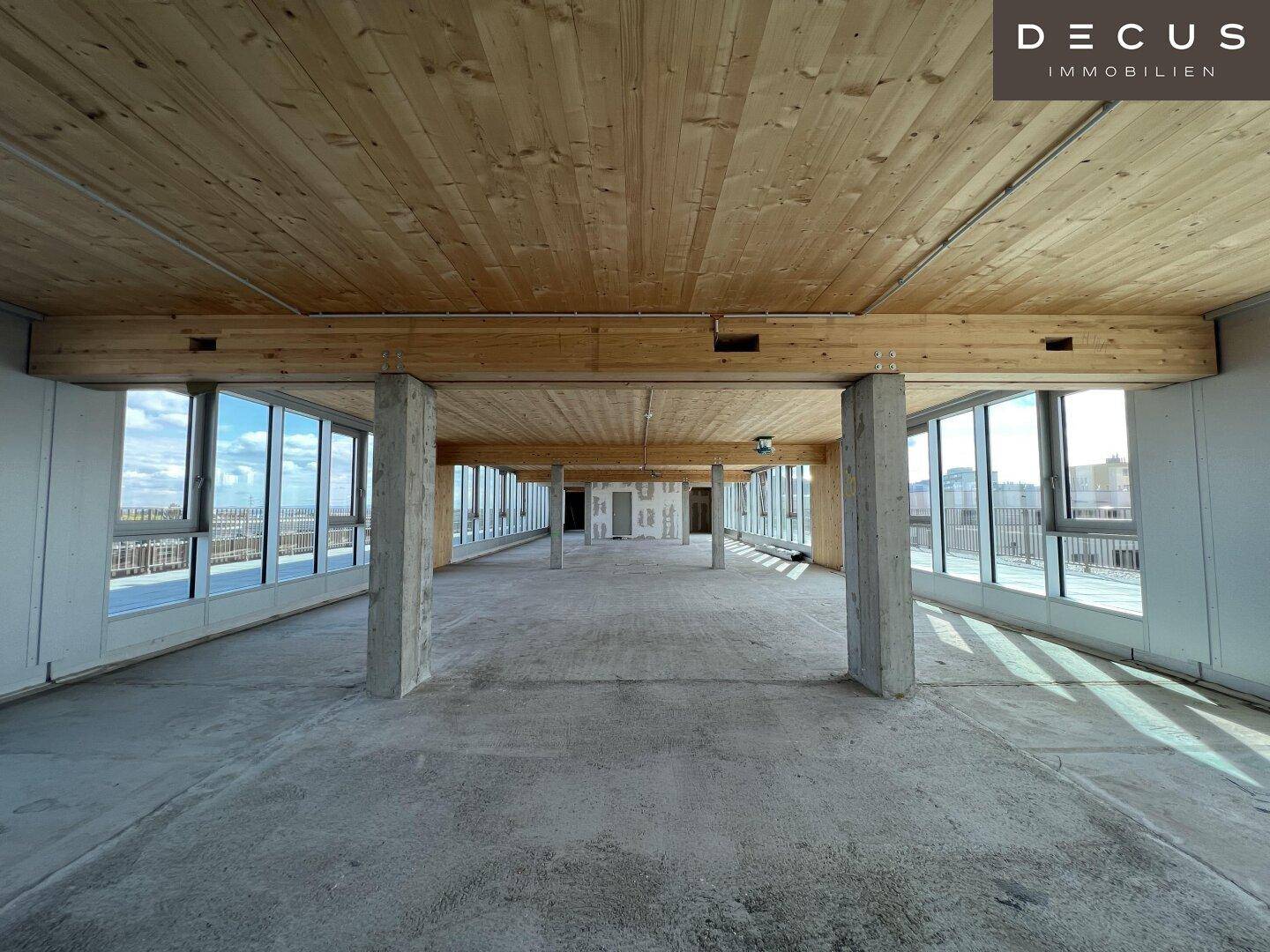 Offener Gewerberaum im Rohbau mit Holzbalkendecke, Betonboden und großen Fenstern.