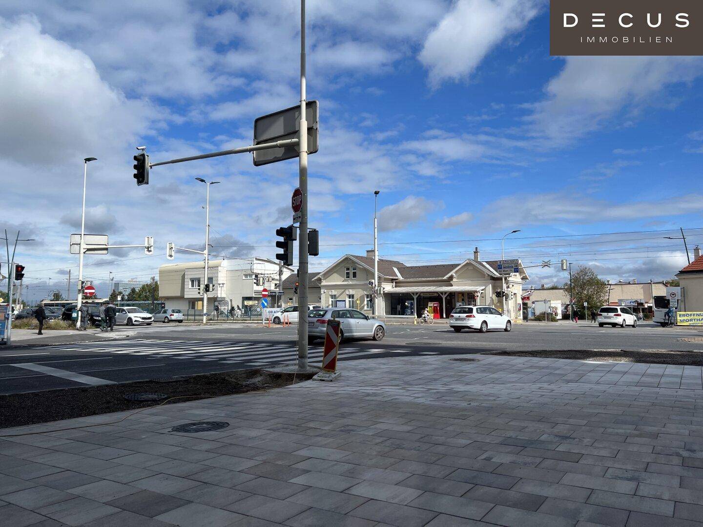 Straßenkreuzung mit Ampeln und einem Bahnhofsgebäude im Hintergrund unter bewölktem Himmel.