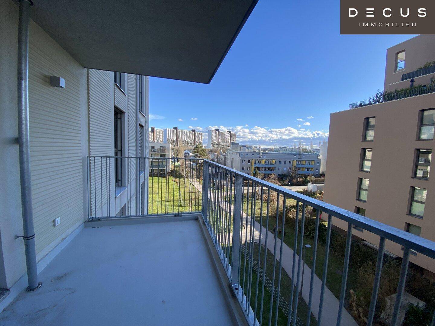 Heller Balkon mit weitem Blick über eine grüne Wiese und moderne Wohngebäude unter blauem Himmel.