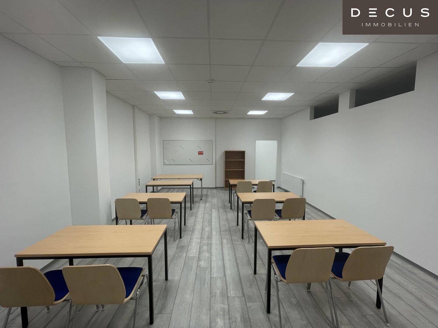 Eingerichteter Konferenz- oder Schulungsraum mit Tischen, Stühlen und Whiteboard, ideal für Besprechungen.
