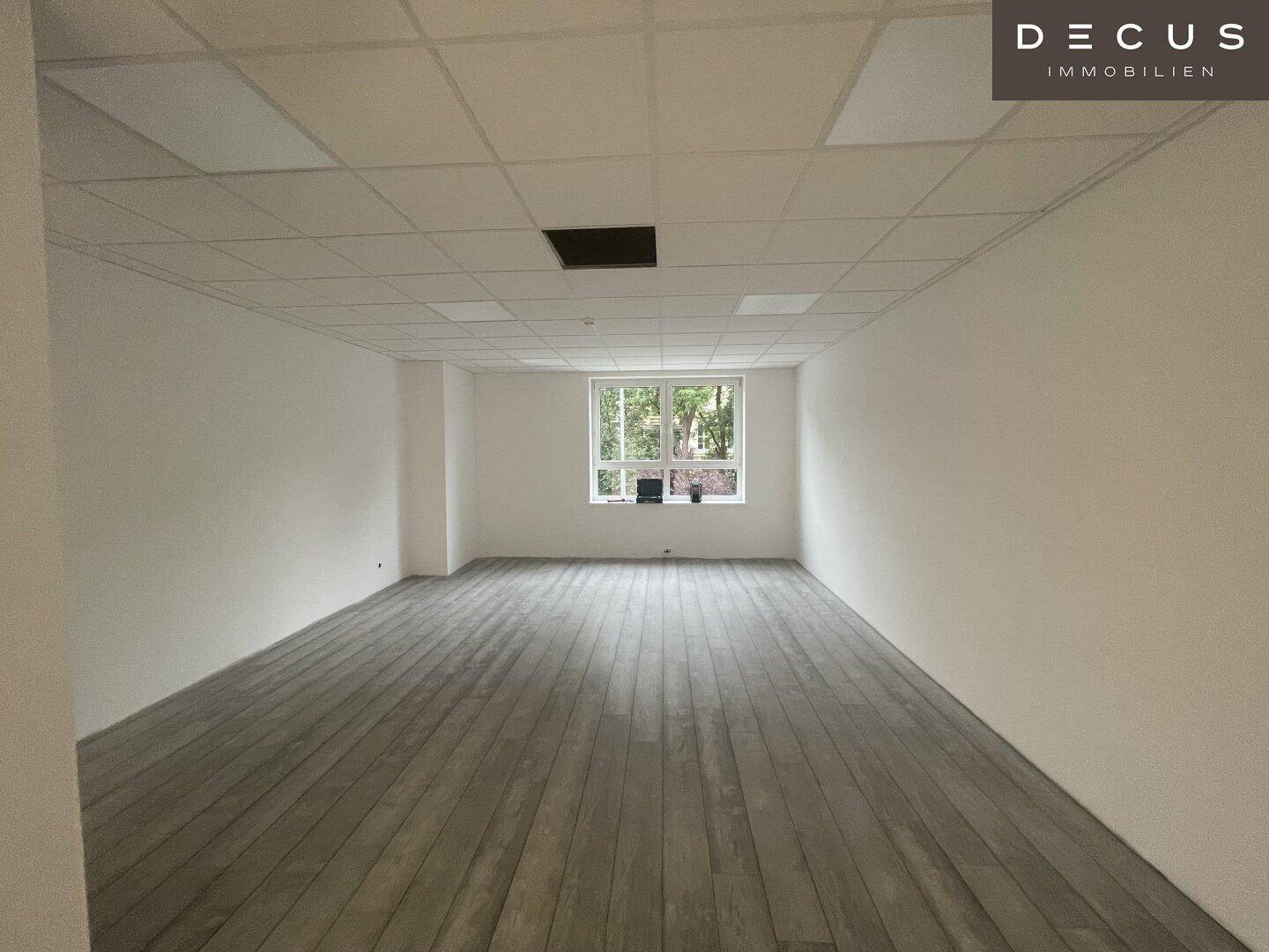Heller Büroraum mit grauem Plankenboden und einem Fenster, das einen Blick ins Grüne bietet.