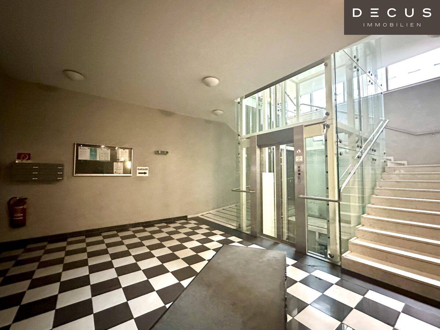 Eingangsbereich mit Schachbrettmuster-Fliesenboden, Treppe und einem modernen Glasaufzug.