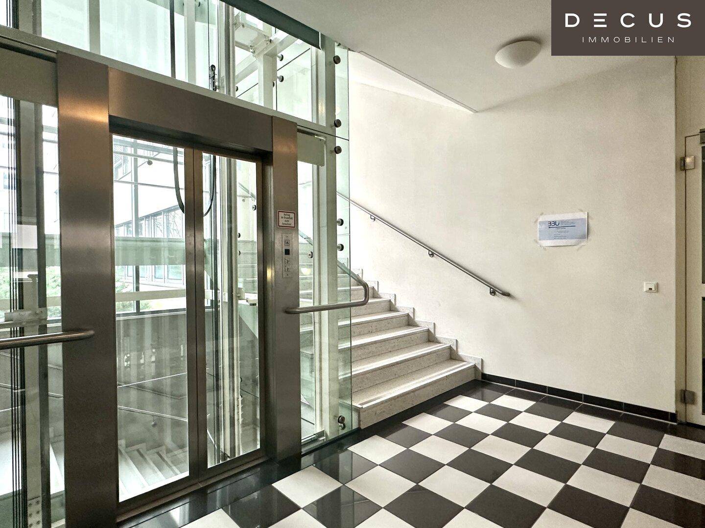 Detailansicht des modernen Glasaufzugs und der Treppe mit Edelstahlgeländer im Eingangsbereich.