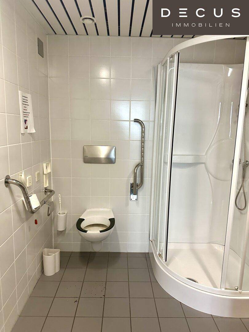 Barrierefreies Badezimmer mit Dusche, WC und Haltegriffen, ausgestattet mit weißen Fliesen.