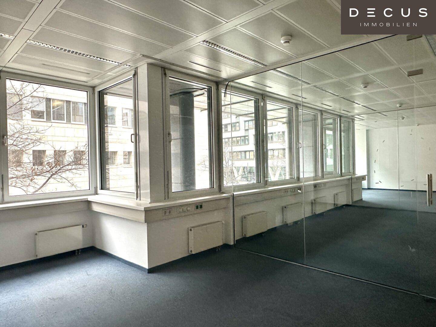 Heller Büroraum mit großen Fenstern, Teppichboden und einer modernen Glaswand, die den Raum unterteilt.