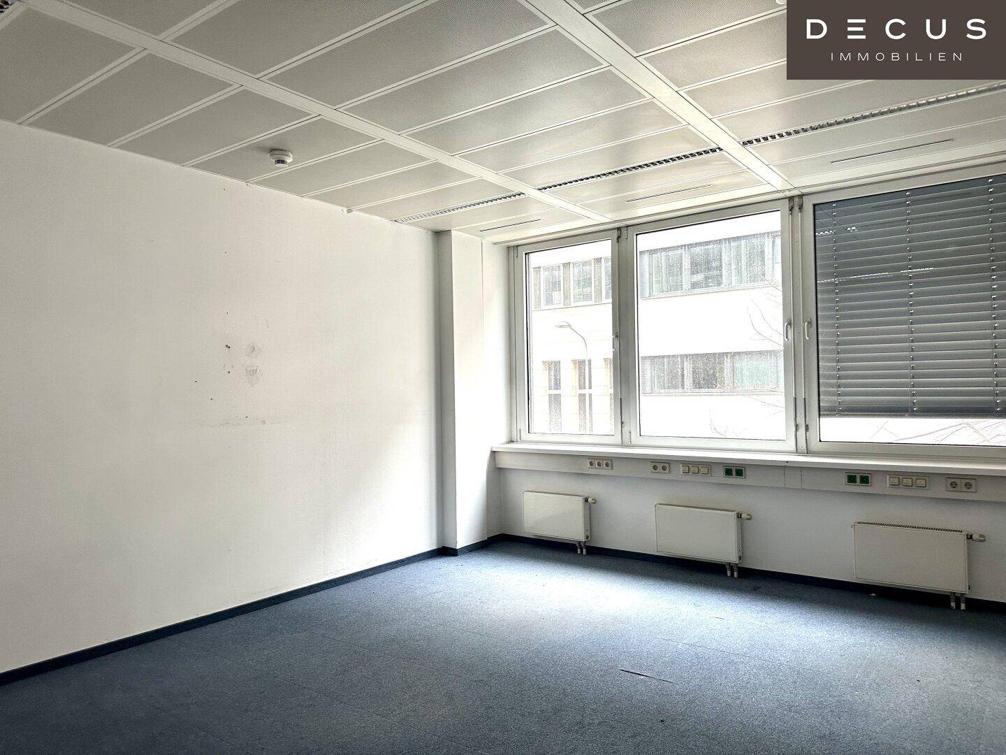 Büroraum mit Teppichboden und Fenstern, die mit Jalousien ausgestattet sind und Tageslicht hereinlassen.