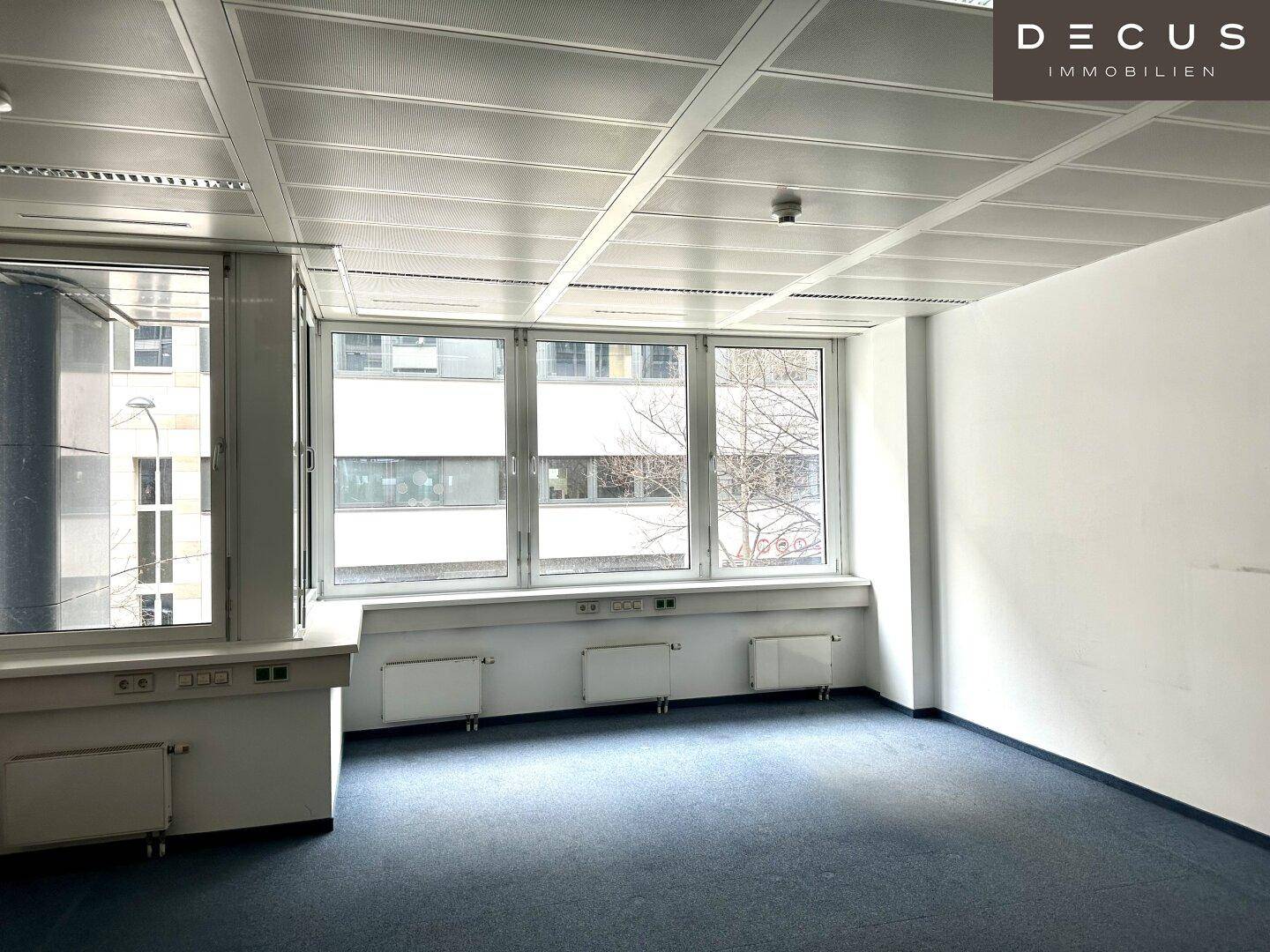 Heller Büroraum mit Teppichboden und großen Fenstern, die eine städtische Aussicht bieten.