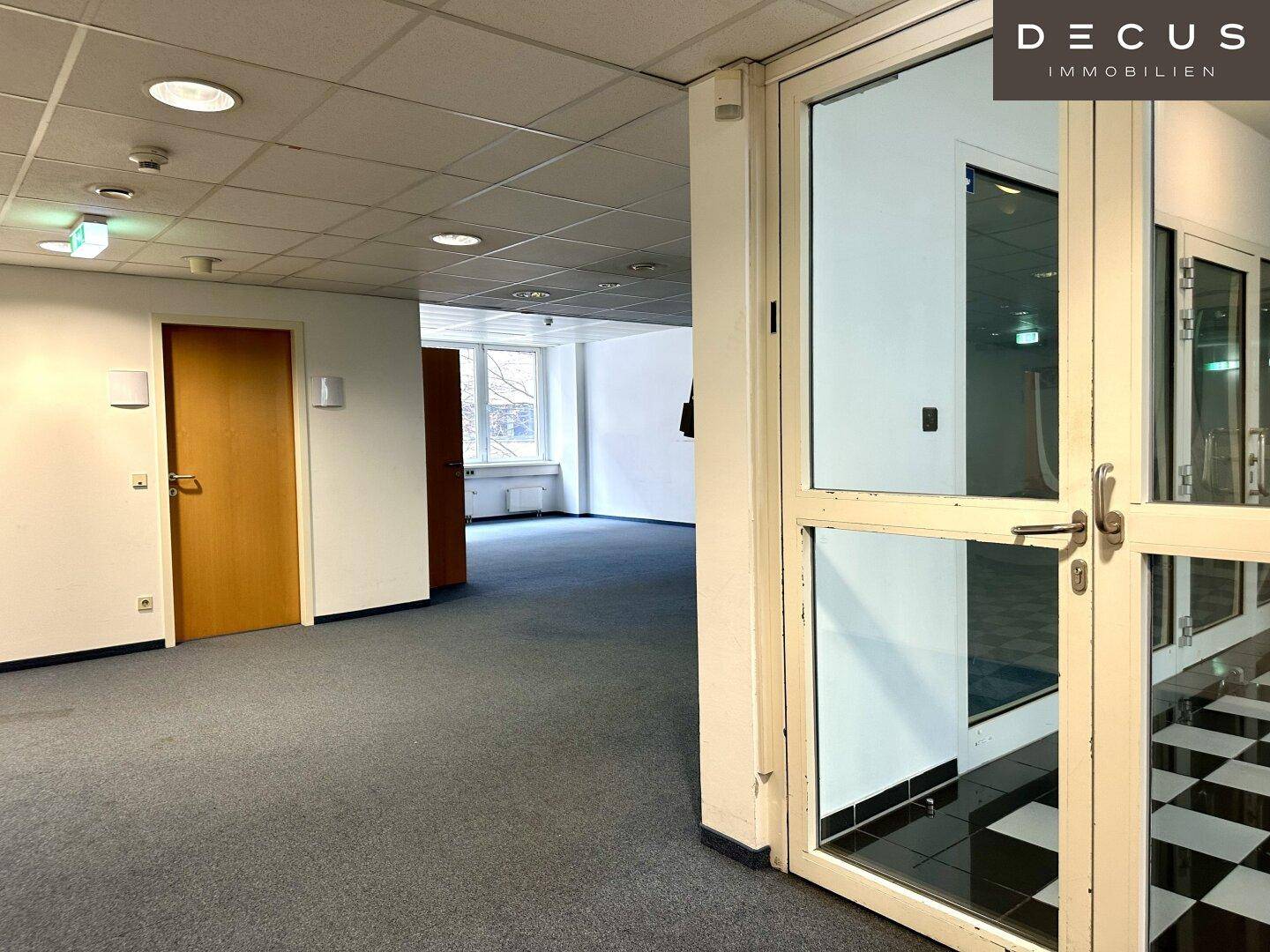Blick durch eine Glastür in einen leeren Büroraum mit Teppichboden und Holztüren.