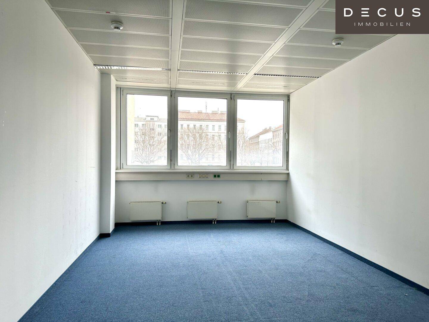 Heller Büroraum mit Teppichboden und drei großen Fenstern, die eine städtische Aussicht bieten.