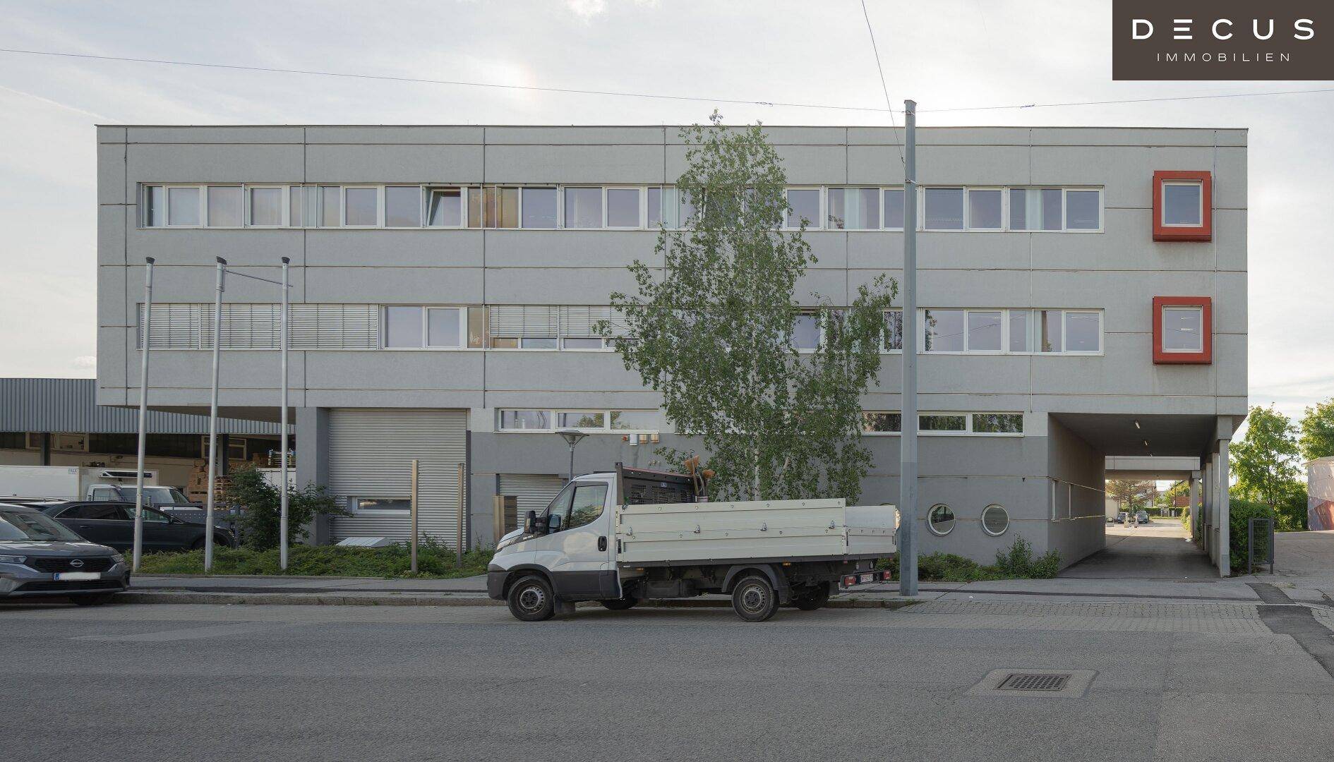 Seitenansicht des Bürogebäudes mit Laderampe und Zufahrt, umgeben von Fahrzeugen und Grünflächen.