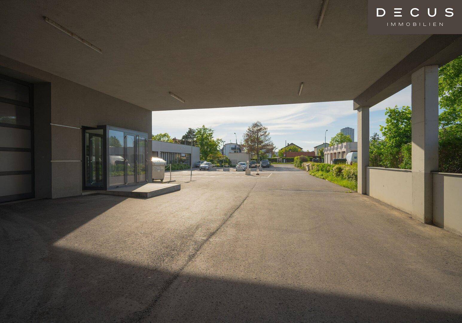 Überdachter Zufahrtsbereich oder Parkplatz mit Blick auf weitere Gebäude und Grünflächen.