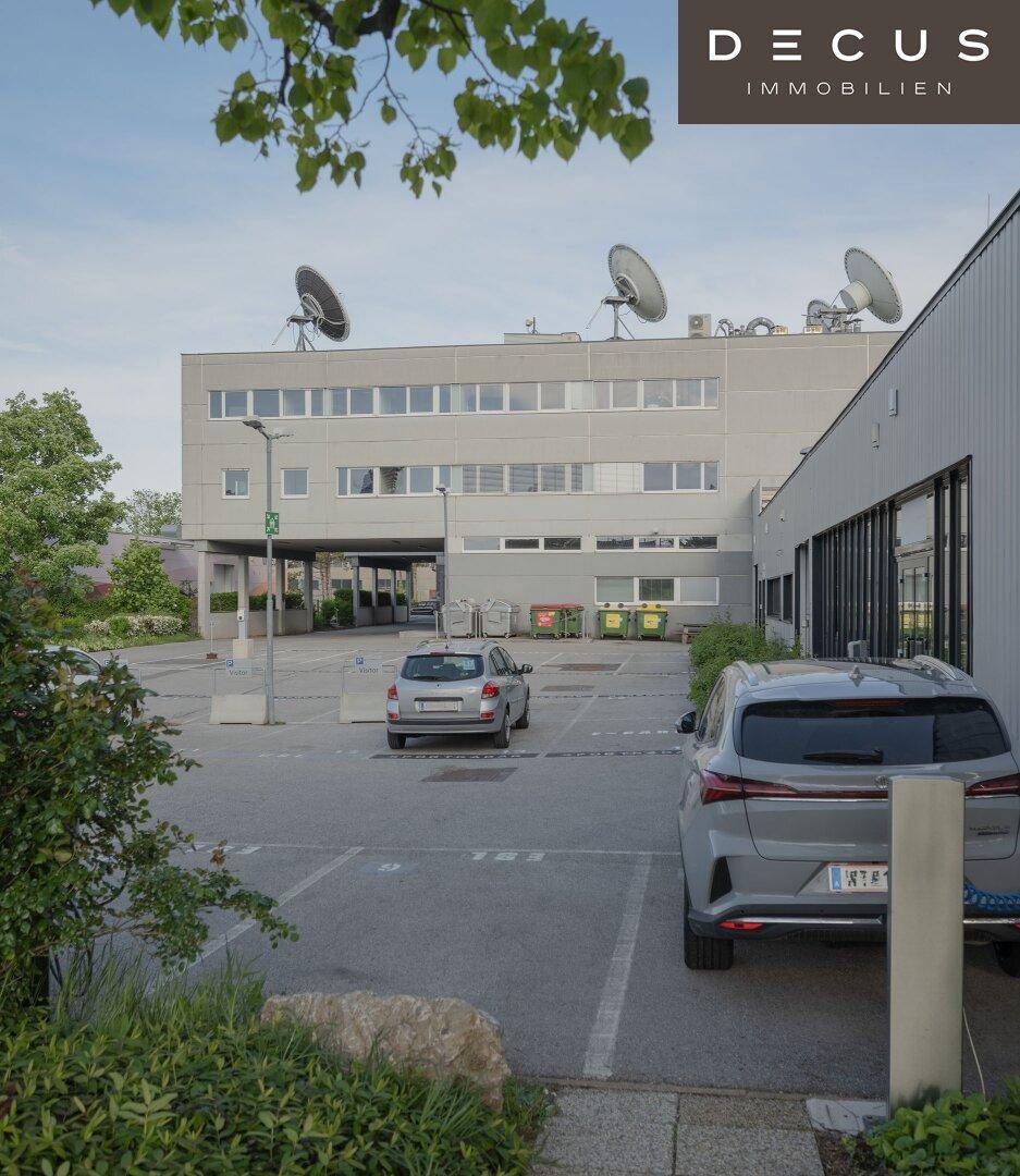 Rückansicht des Bürogebäudes mit Parkplatz, Mülltonnen und Satellitenschüsseln auf dem Dach.
