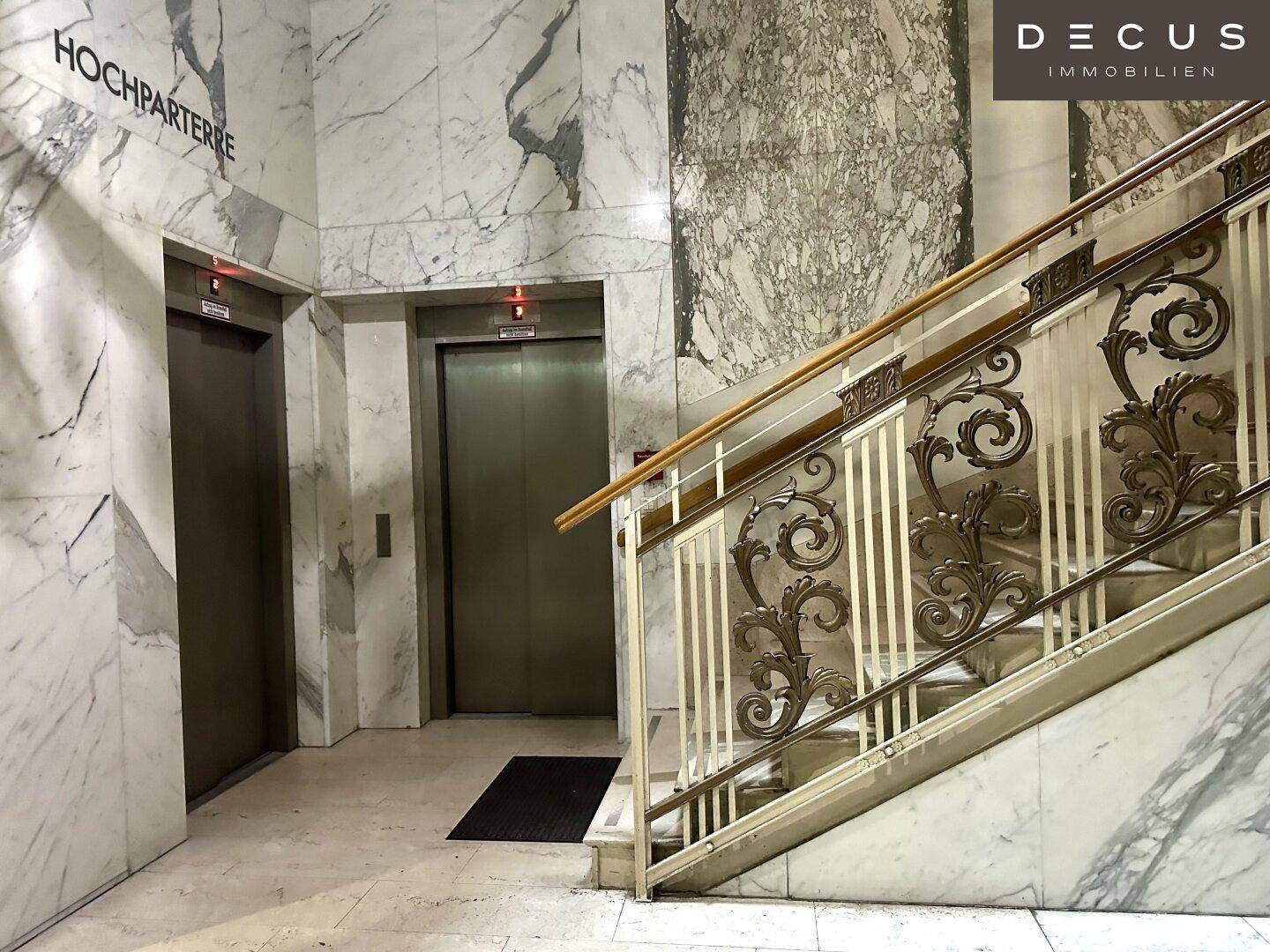 Heller Empfangsbereich mit zwei Aufzügen und einer kunstvollen Treppe, umgeben von Marmor.