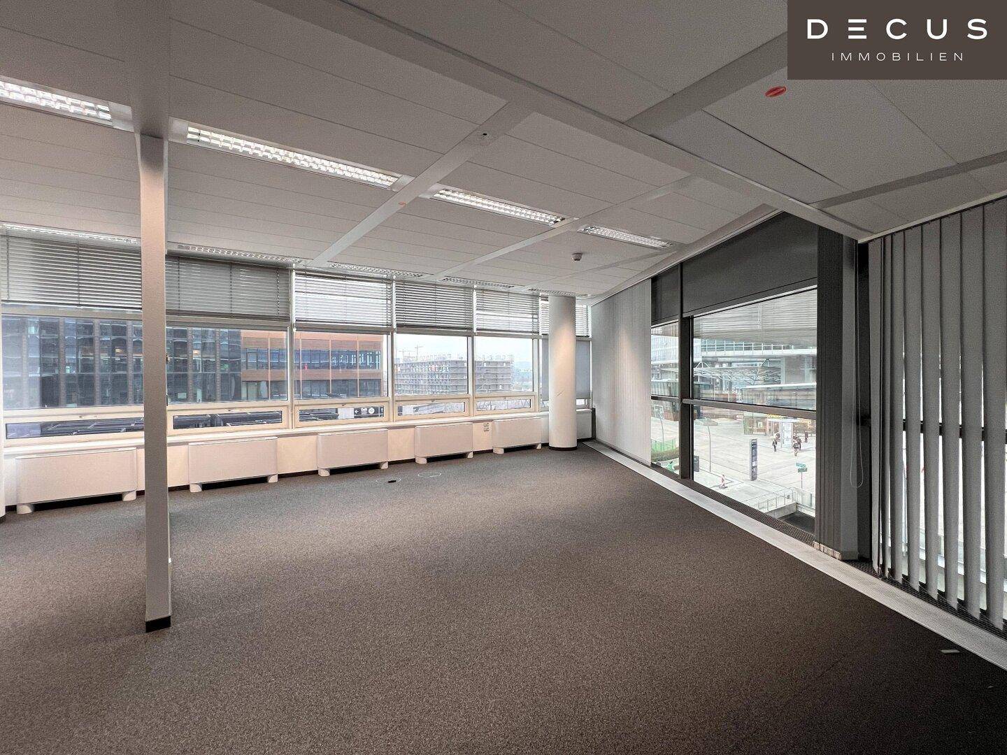 Leeres Büro mit Teppichboden und breiter Fensterfront, die einen urbanen Ausblick bietet.