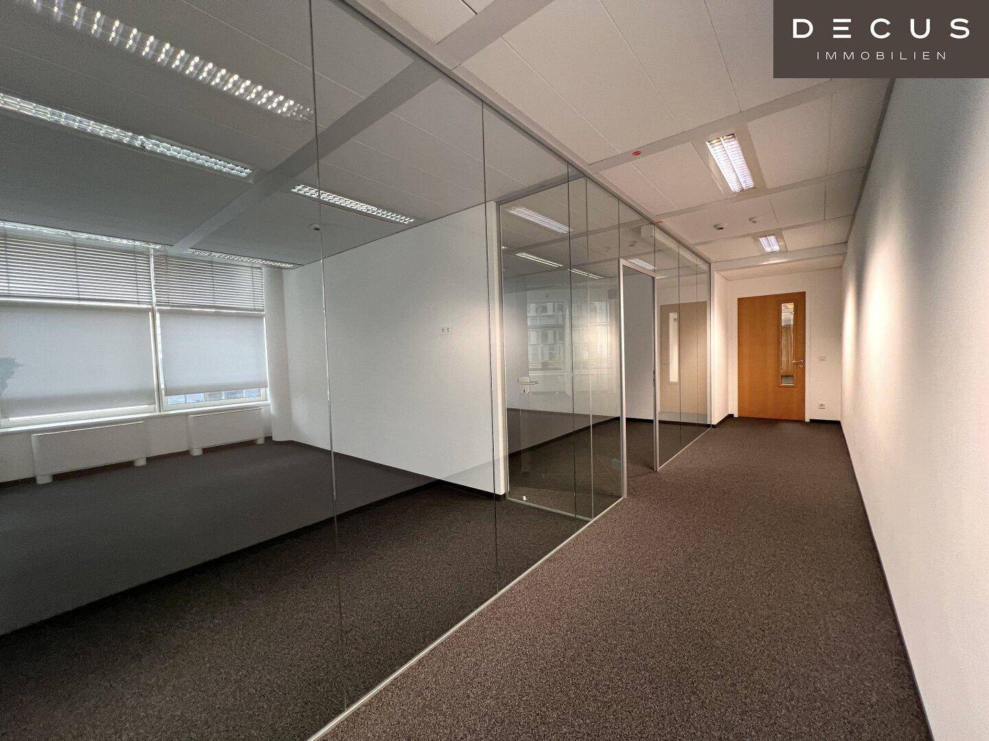 Büroflur mit Glaswänden und Teppichboden, der zu weiteren Büroräumen führt.