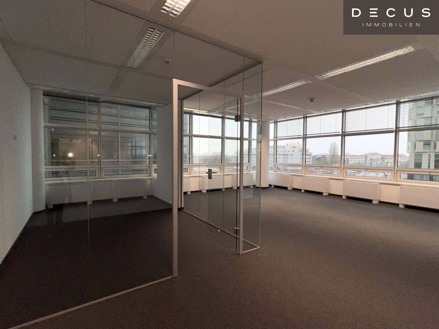 Großes Eckbüro mit Teppichboden und raumhohen Fenstern, die viel Tageslicht hereinlassen.