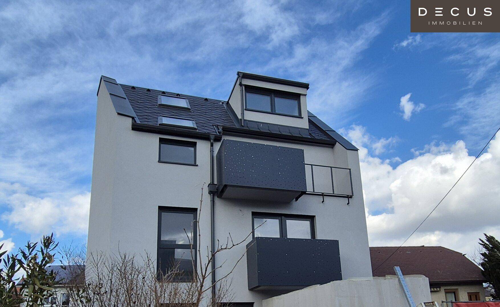 Moderne Fassade eines Mehrfamilienhauses mit Balkonen und Dachfenstern unter blauem Himmel.