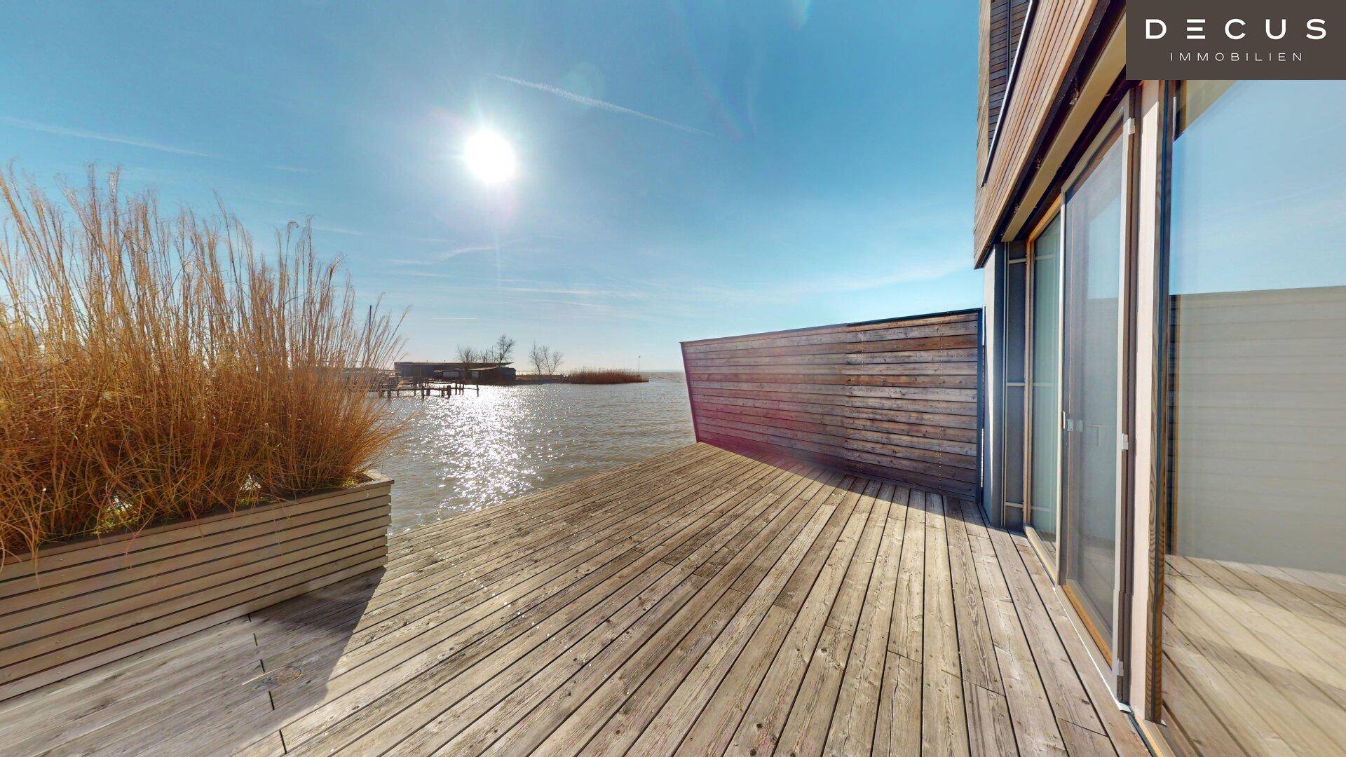 Weitläufige Holzterrasse mit Blick auf den See und Schilfbepflanzung am Ufer.