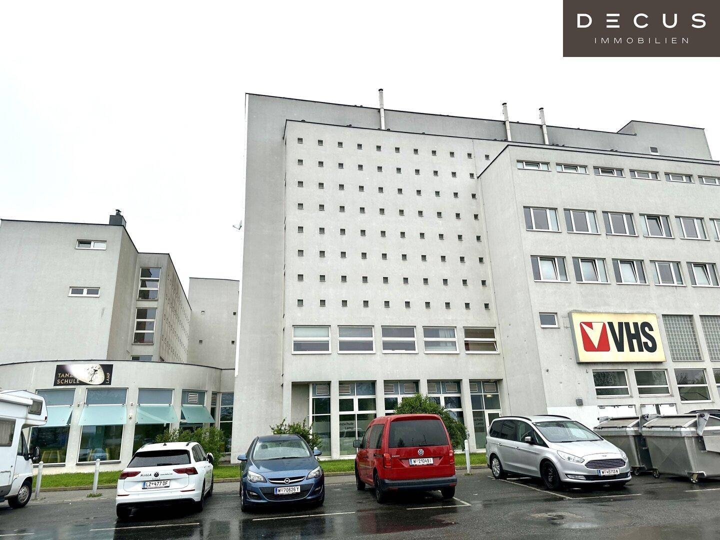 Mehrstöckiges Gebäude mit Parkplätzen davor und verschiedenen Geschäften im Erdgeschoss.