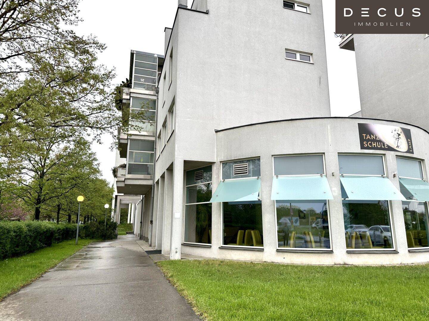 Moderne Gebäudefassade mit runden Elementen und großen Fenstern, umgeben von Grünflächen.