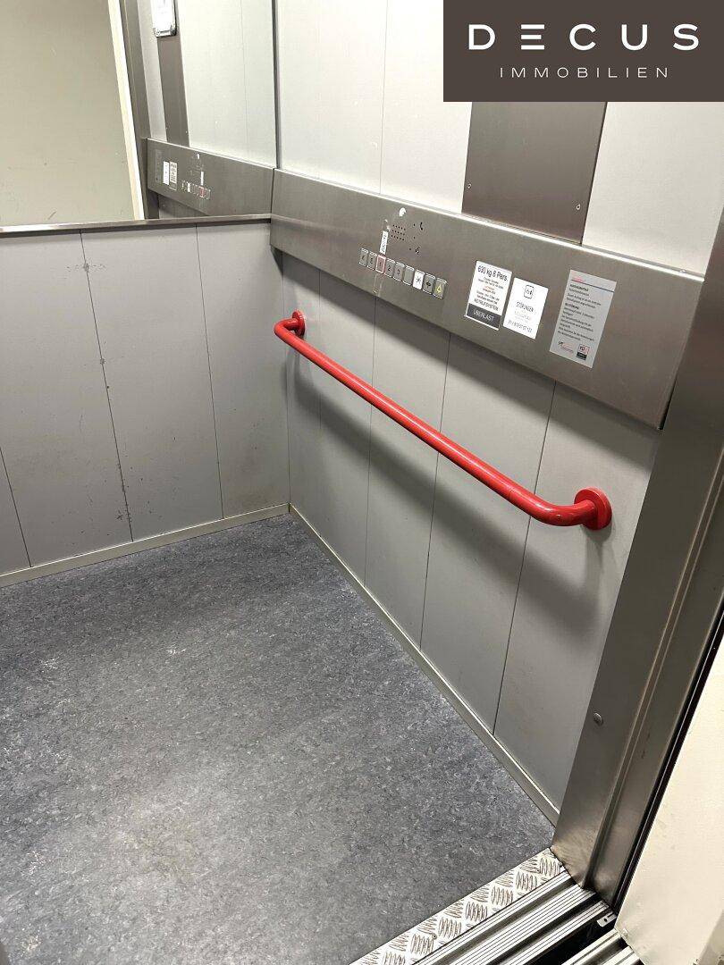 Innenansicht eines Aufzugs mit rotem Haltegriff und Metallverkleidung, barrierefrei.