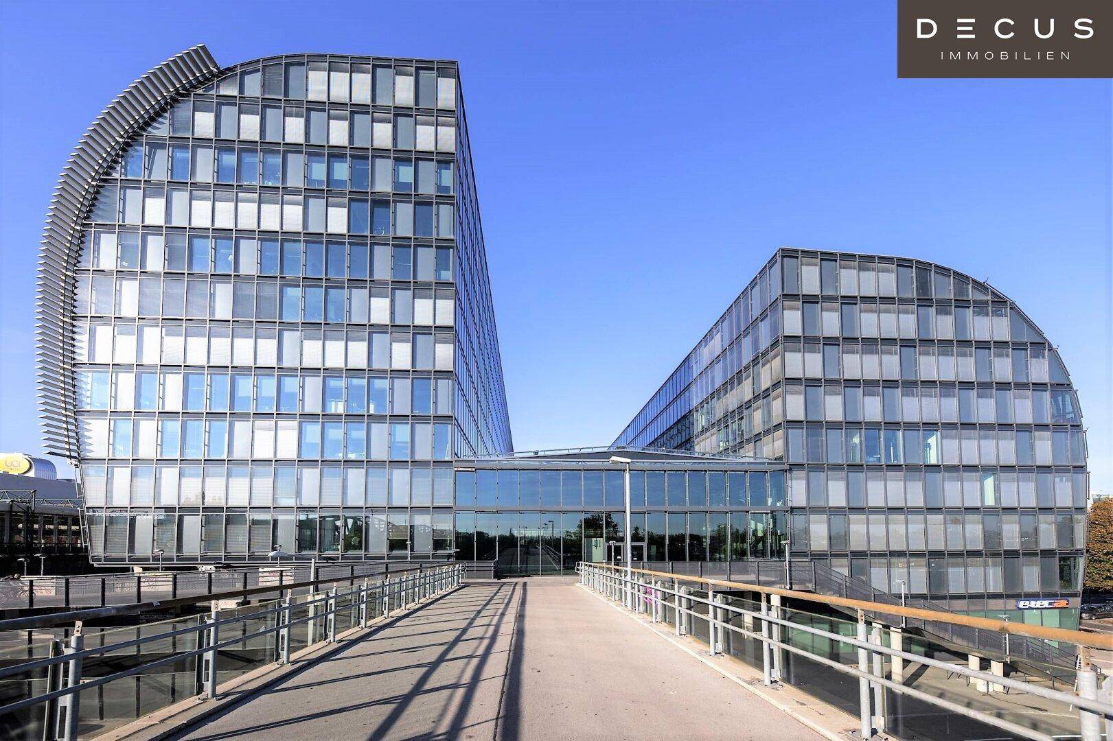 Moderne Bürogebäude mit geschwungener Glasfassade und einer erhöhten Fußgängerbrücke unter blauem Himmel.