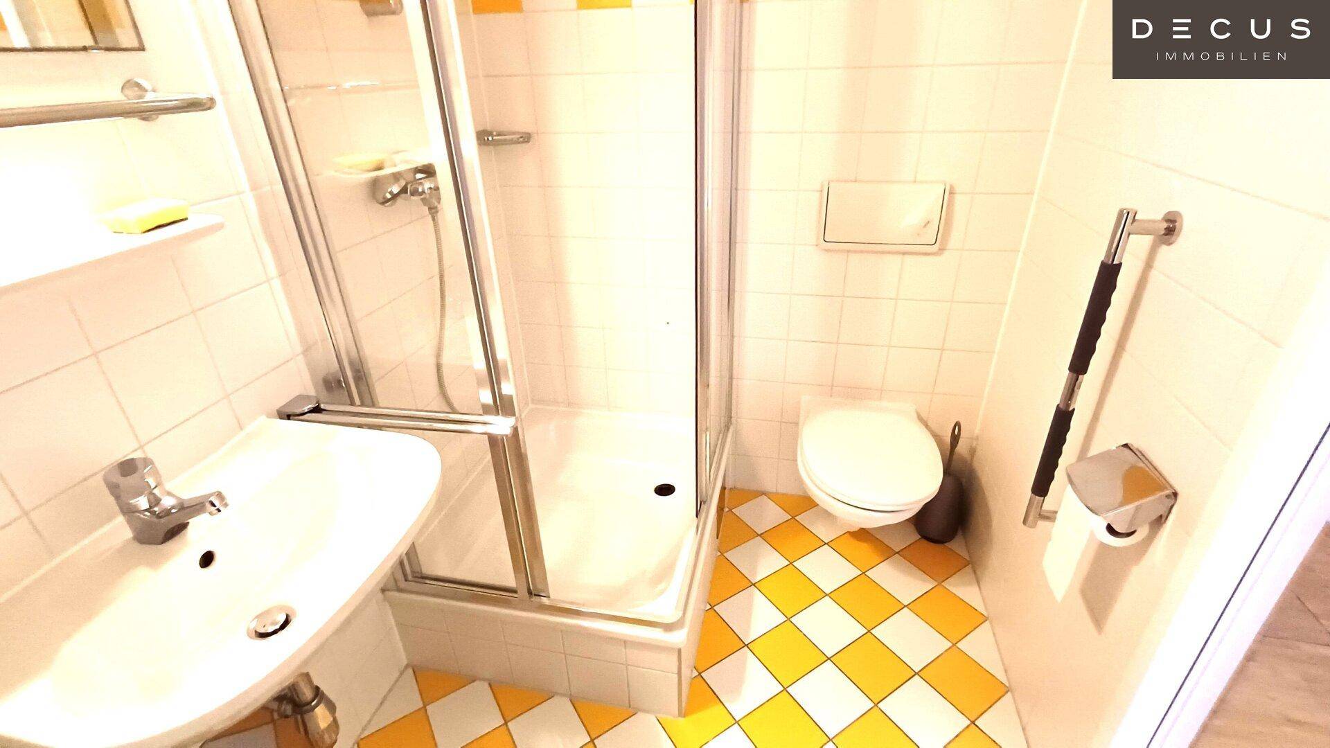 Badezimmer mit Duschkabine, WC und gelb-weiß karierten Fliesen am Boden sowie Haltegriffen.