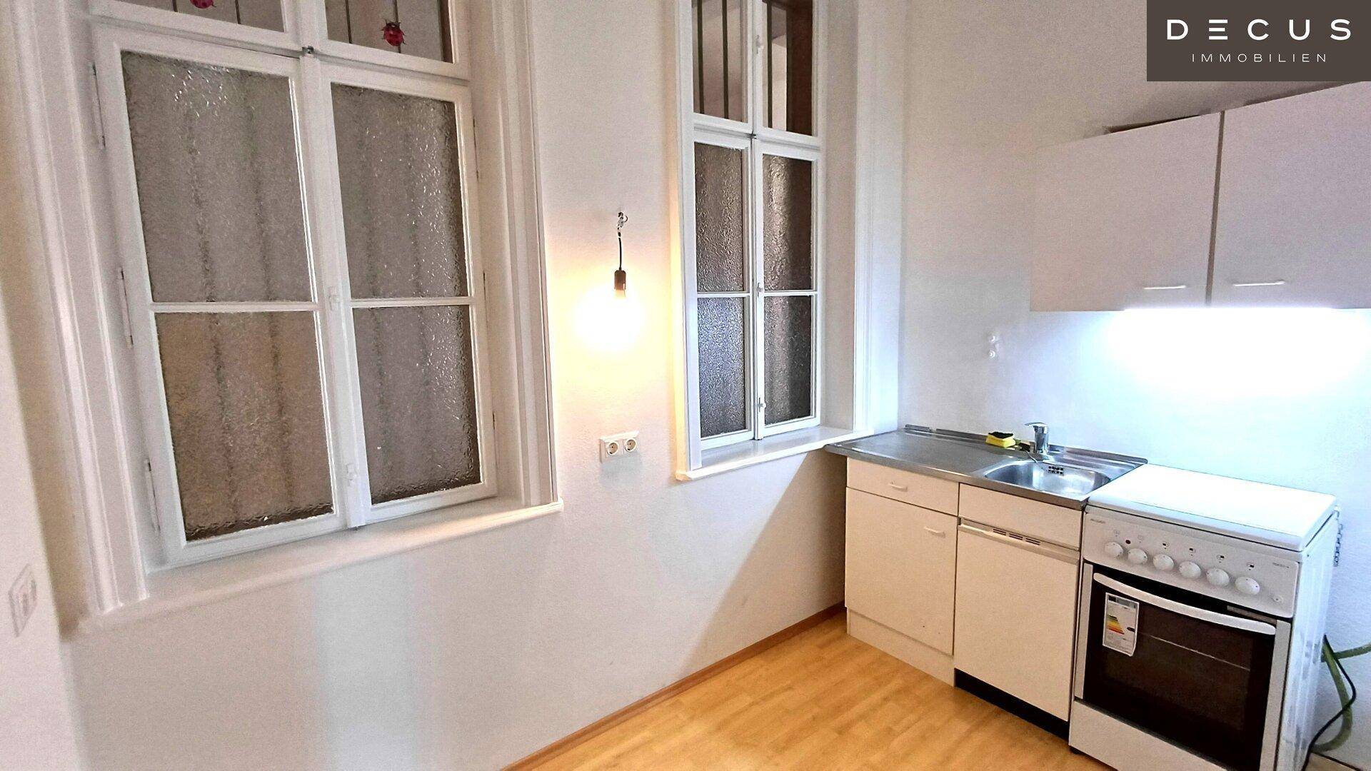 Kleine Küchenzeile mit Spüle, Herd und Oberschränken unter zwei Fenstern in einem hellen Raum.