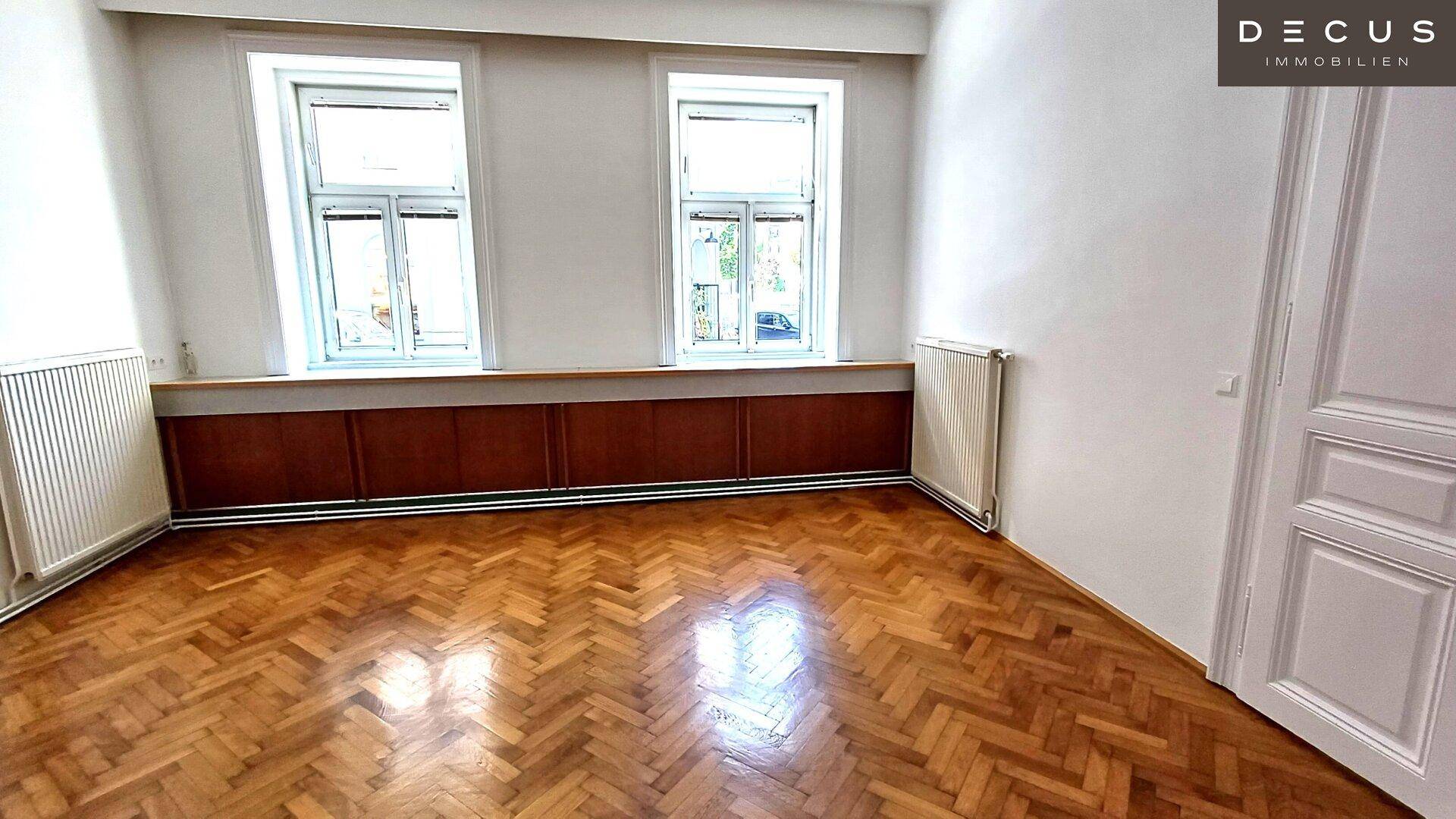 Großes Zimmer mit Fischgrätparkett, zwei Fenstern und Heizkörpern unter der Fensterbank.