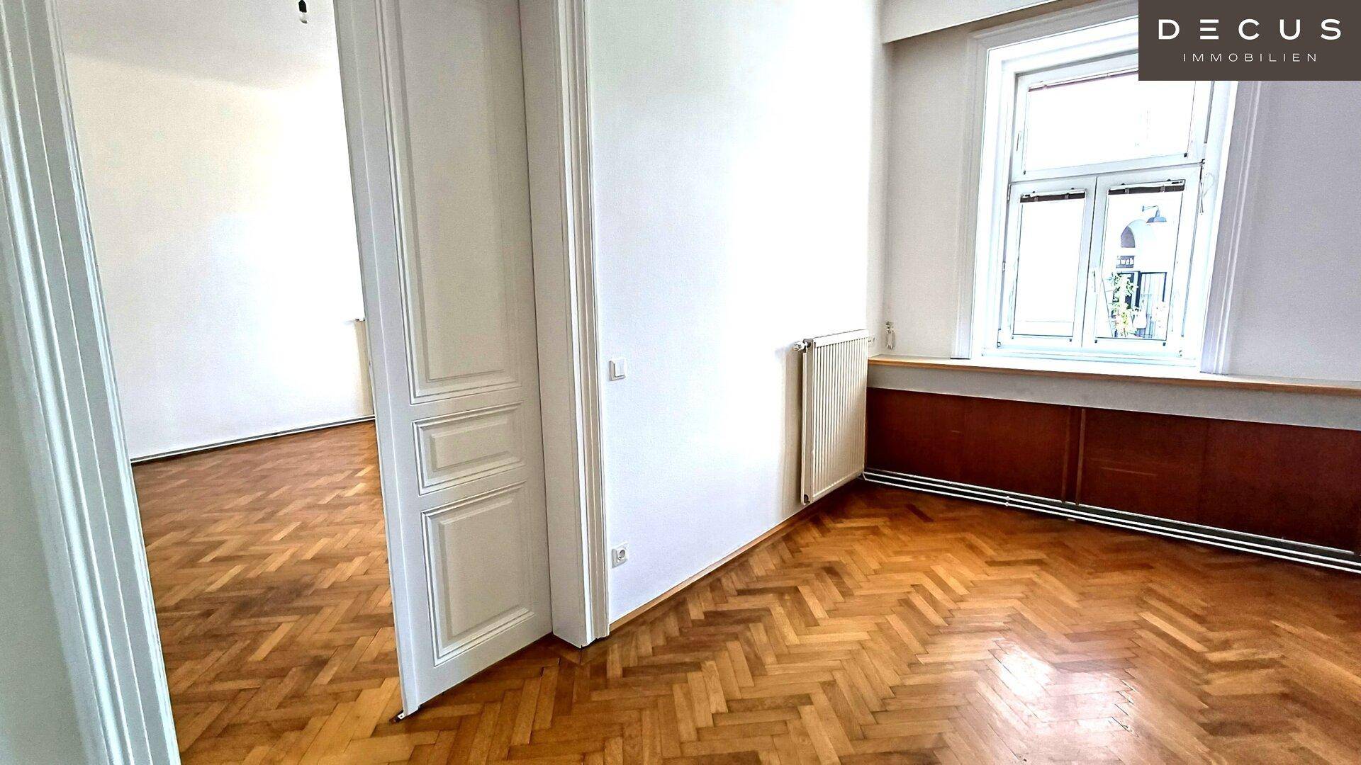 Helles Zimmer mit Fischgrätparkettboden, Fenster und einer geöffneten Tür zu einem weiteren Raum.