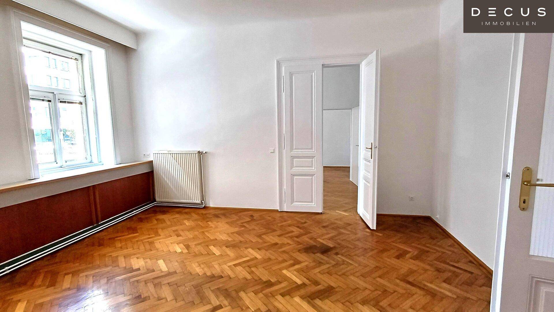Geräumiges Zimmer mit Fischgrätparkett, Heizkörper und einem Fenster mit Holzeinfassung.