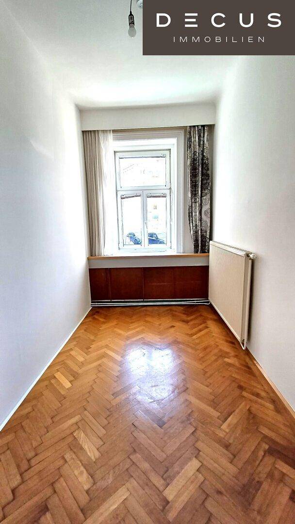 Langes, schmales Zimmer mit Fischgrätparkett, einem Fenster und Vorhängen an der Wand.