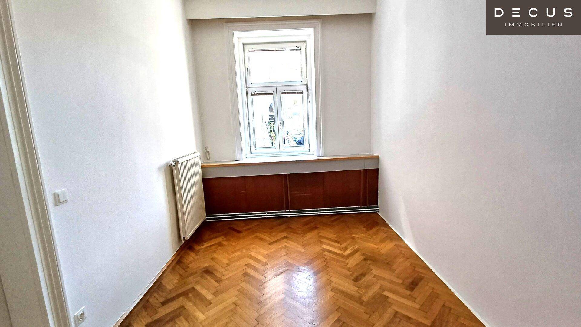 Kleineres Zimmer mit Fischgrätparkett, einem Fenster und einem Heizkörper an der Wand.